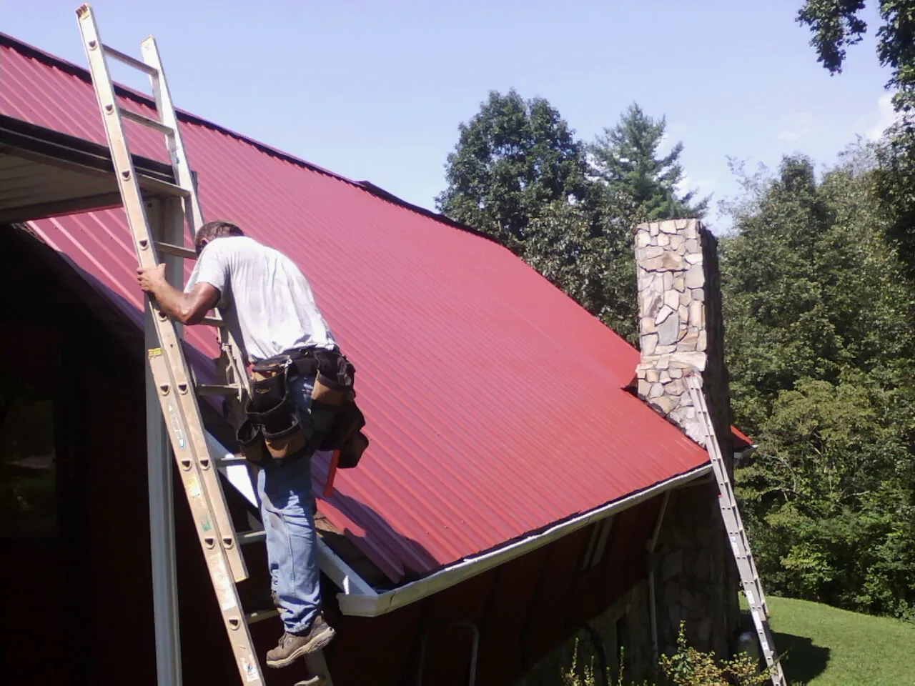 Roofer on a ladder beside a red metal roof and stone chimney — Soffit & Fascia Repair by Roofing Brothers in Converse